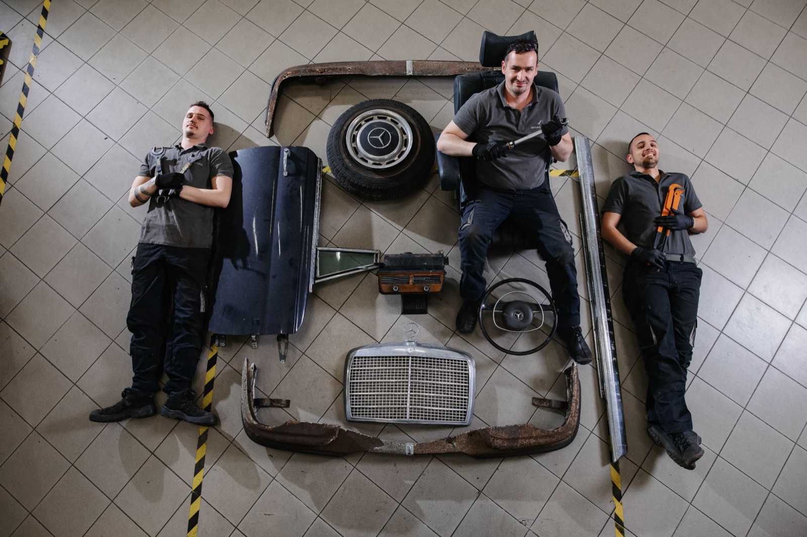 Three mechanics lying on the workshop floor around the parts of a Mercedes-Benz laid out in the shape of a car, holding their tools