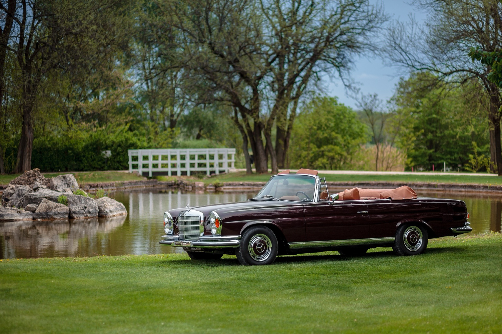 Maroon Mercedes-Benz W111 convertible on a lawn by a lake
